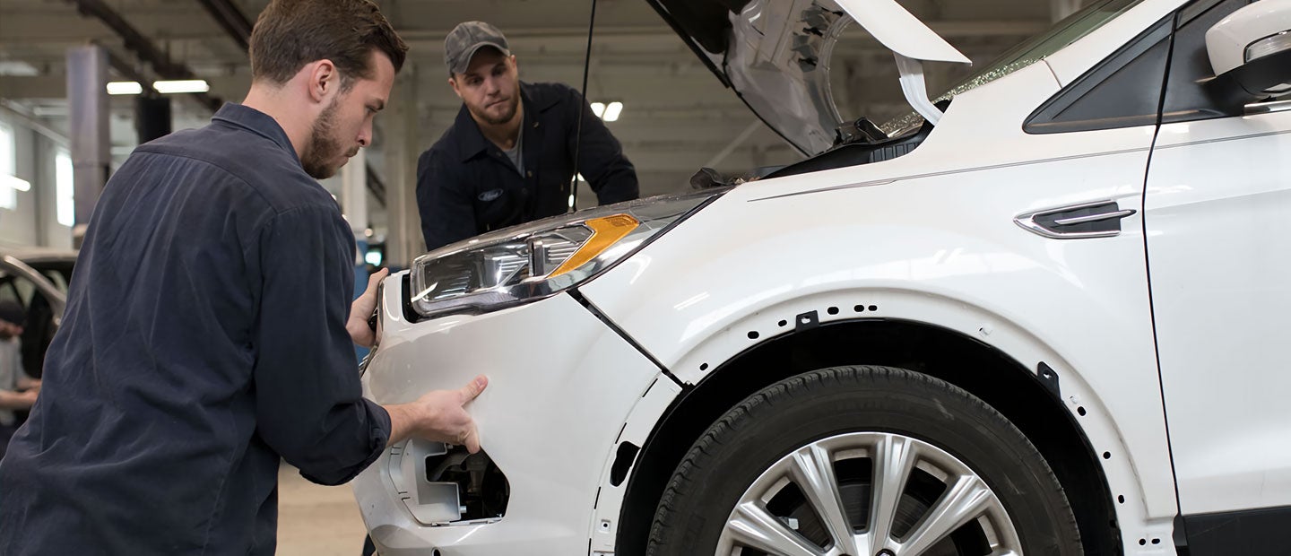 Two mechanics work on a white car, one adjusting the front bumper, the other looking under the open hood.