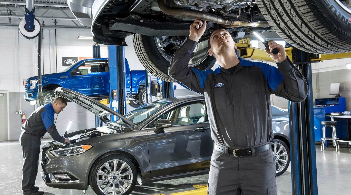 Two Ford mechanics working on cars in a service bay; one inspects a car on a lift, another a car's engine.