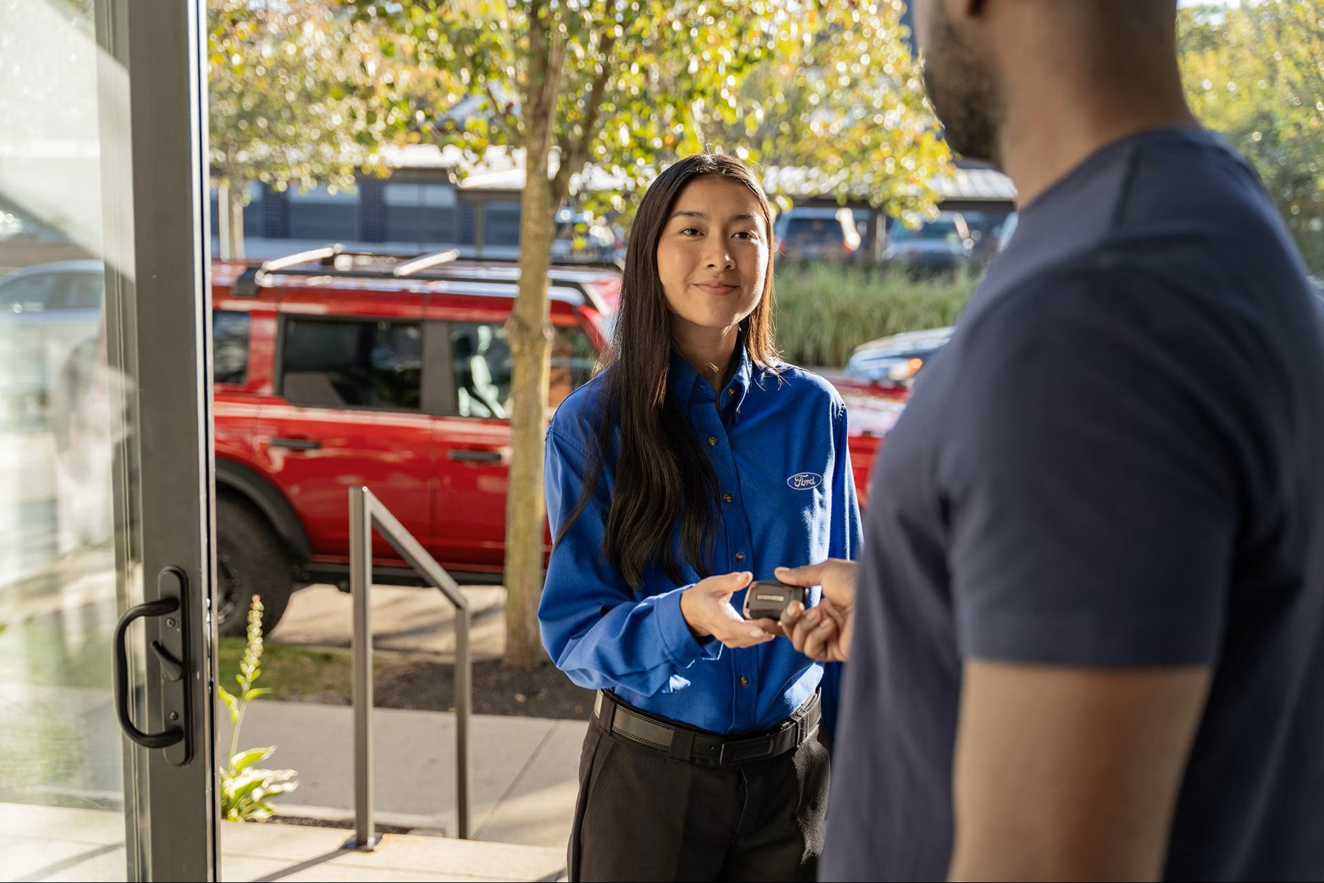 Woman in Ford shirt handing car keys to a man, with a red Ford Bronco visible in the background.