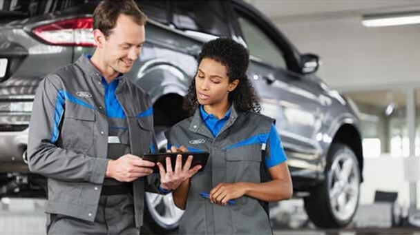 Two mechanics in Ford uniforms, a man and a woman, discuss information on a tablet in a service center with a car.