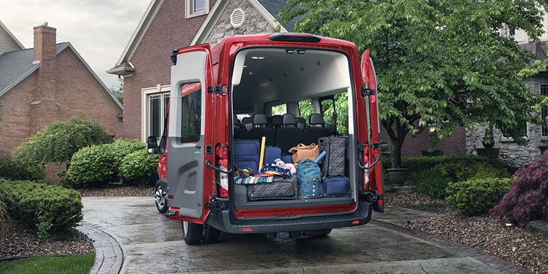 Red passenger van with its back doors open, revealing a cargo area full of luggage, parked in a wet driveway.