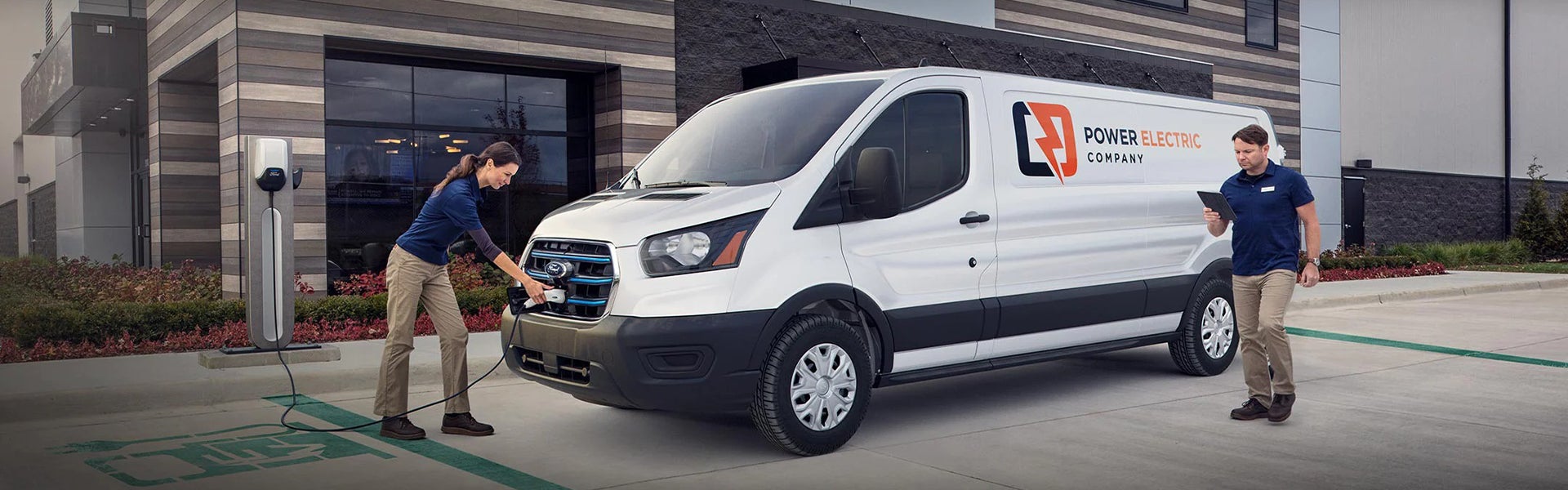 A woman plugs a white Ford Electric van from "Power Electric Company" into a charging station as a man uses a tablet.
