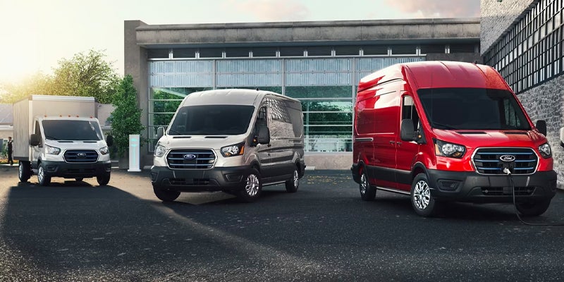 Three Ford Transit vans: a white box truck, a silver cargo van, and a red electric cargo van charging at a station.