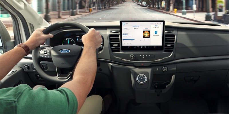 Person driving a Ford van, hands on the steering wheel, with a large touchscreen displaying music in the dashboard.