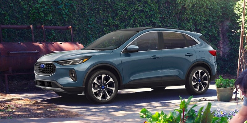 A blue-green Ford Escape parked on a paved surface next to an outdoor grill and lush green foliage.