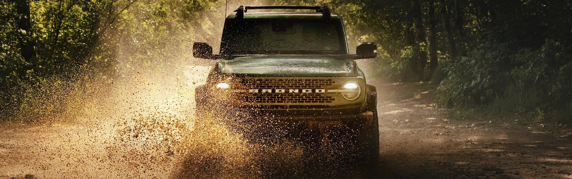 A green Ford Bronco drives on a muddy forest road, splashing mud and water forward, with headlights on.