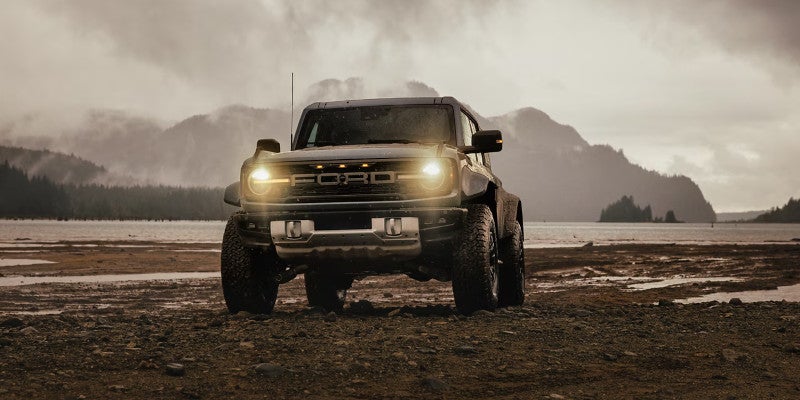 Dark Ford Bronco with headlights on, parked on a muddy shore with a lake and misty, forested mountains in the background.