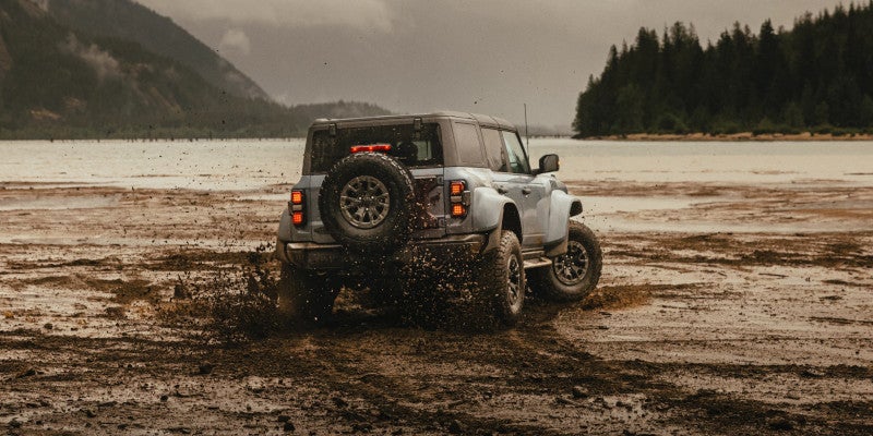 Grey SUV splashing mud as it drives through a muddy landscape next to a lake and mountains under a cloudy sky.