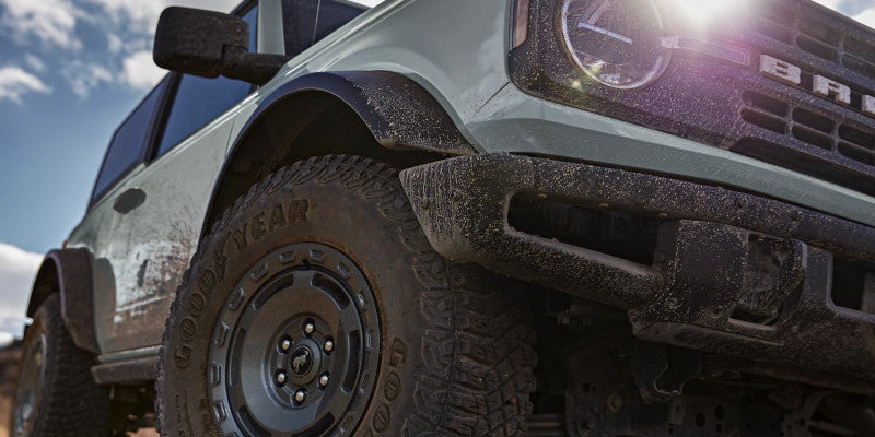 Muddy, light green Ford Bronco from a low angle, showing front wheel with Goodyear tire and dirty grille.