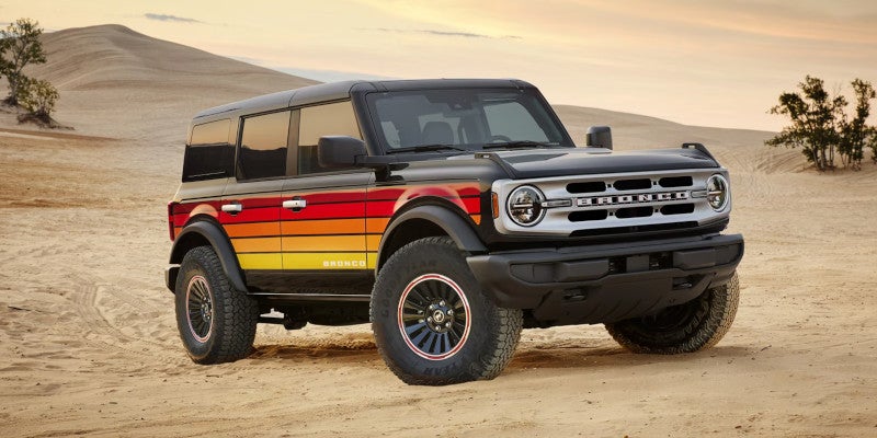 Black Ford Bronco with retro red, orange, and yellow stripes on a sand dune at sunset.