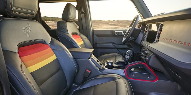 The interior of a Ford F-150 with a steering wheel, digital dashboard, and large infotainment screen displaying a mapCar interior with black leather seats, colorful striped inserts, Bronco logo, and a visible dashboard..