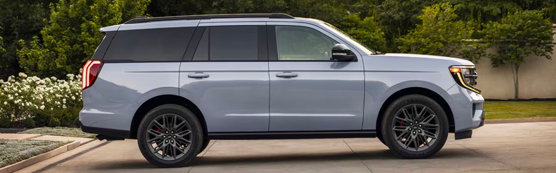 Light gray SUV parked on a paved driveway, with green trees and white flowering bushes in the background.