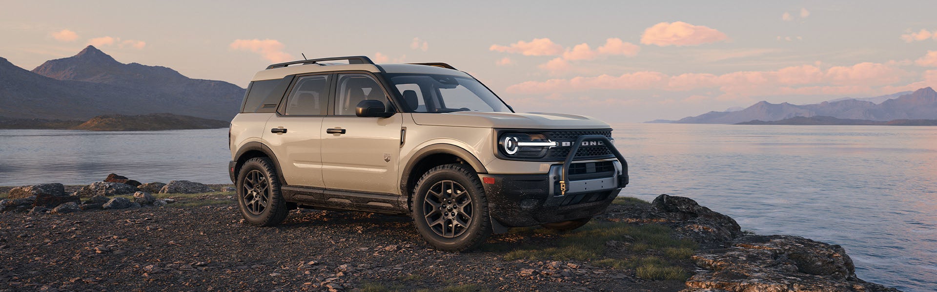 Tan Ford Bronco Sport SUV parked on a rocky shore by a lake, with mountains and a sunset sky in the background.