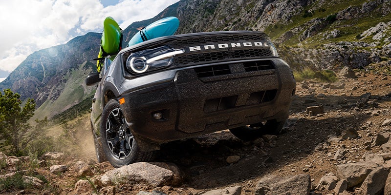 Gray Ford Bronco with two kayaks on the roof drives up a rocky mountain trail under a bright sky.