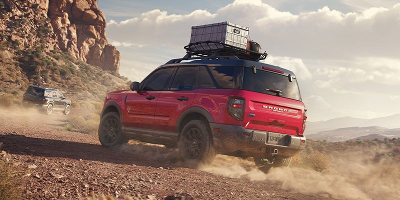 Red Ford Bronco Sport with roof cargo box kicks up dust on a desert dirt road, another Bronco Sport follows behind.