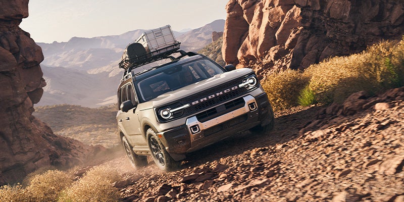 Light tan Ford Bronco with roof rack driving down a rocky desert hill with mountains in the background.