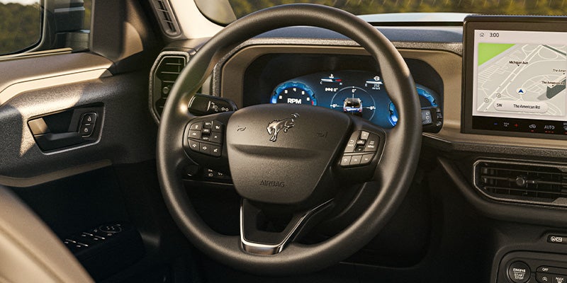 Car interior showing a steering wheel with a Bronco logo, digital instrument cluster, and a large navigation screen.