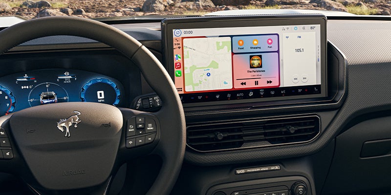 Car interior showing a steering wheel with a Bronco logo and a large infotainment screen displaying Apple CarPlay with maps and music.
