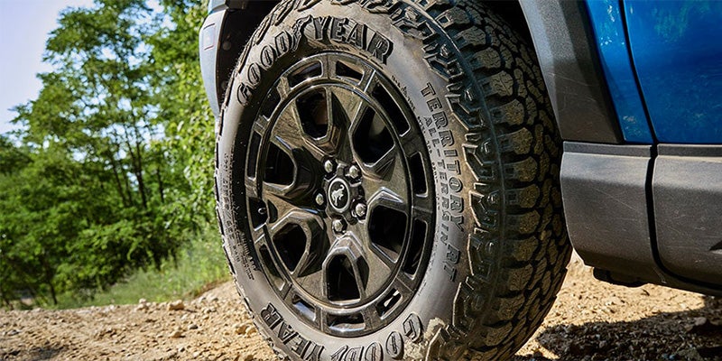 Close-up of a vehicle's dark gray wheel with a Bronco logo and a Goodyear Territory all-terrain tire on a dirt trail.