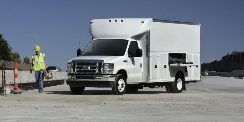 White Ford service truck with an open utility compartment door on a roadway, with a construction worker walking nearby.