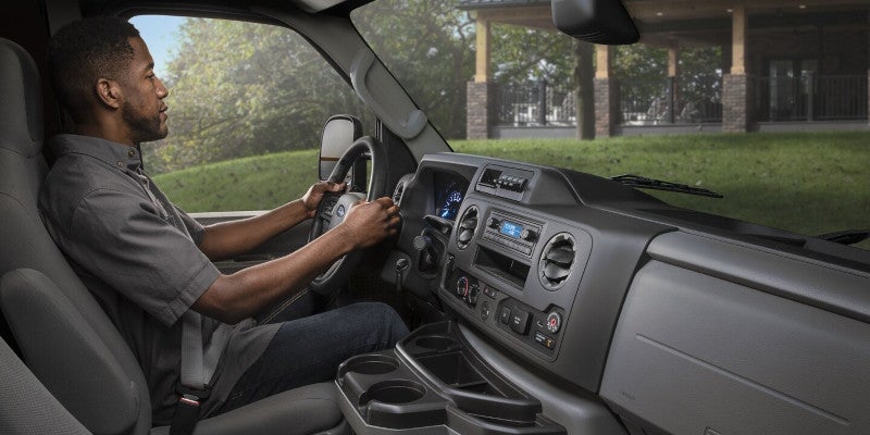 Man driving a commercial van, seen from the side, looking forward. Dashboard, steering wheel, and front window view are visible.