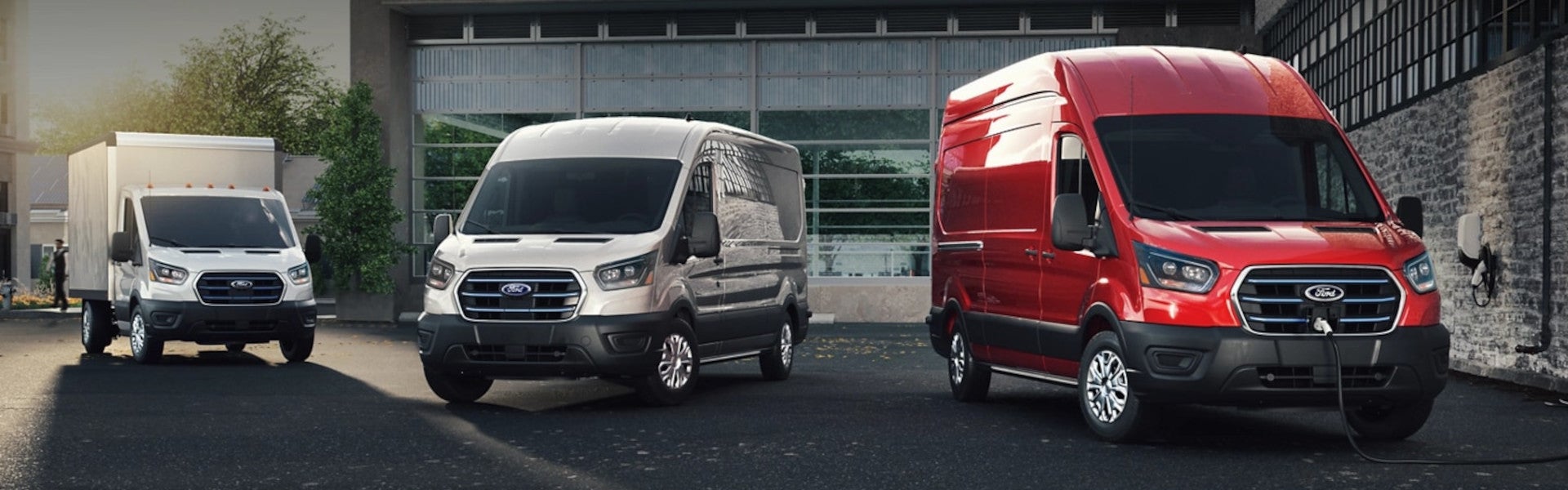 Three electric Ford vans: a white box truck, a silver cargo van, and a red cargo van charging.