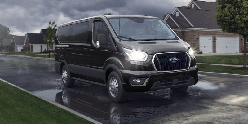 Black Ford Transit van driving on a wet suburban street in the rain with bright headlights and reflections.
