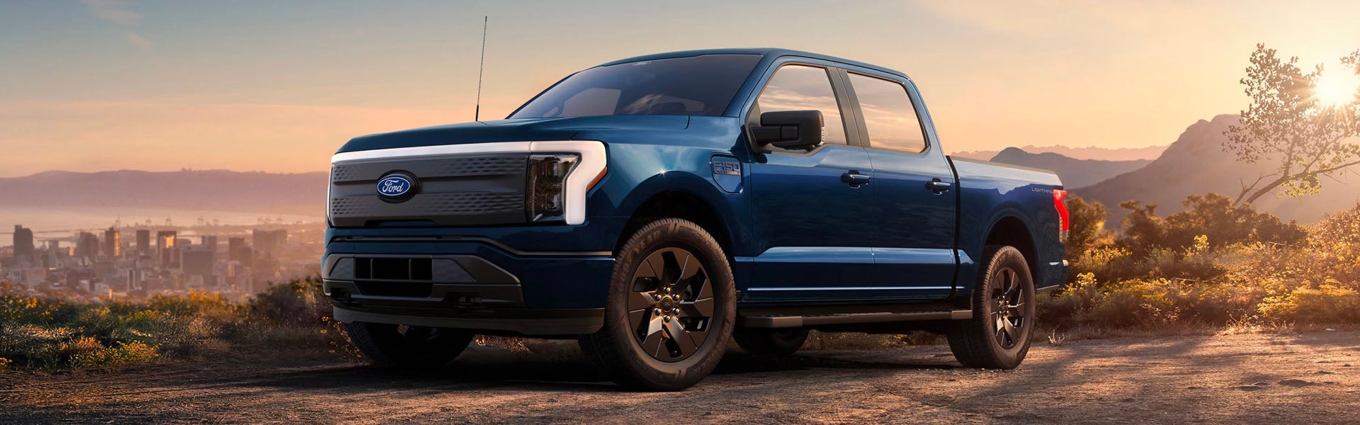 Blue Ford F-150 Lightning electric truck on a dirt road, with a city skyline and mountains in the background at sunset.