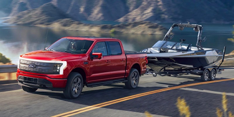 Red Ford F-150 Lightning truck towing a boat on a trailer, driving on a road next to a lake with mountains.
