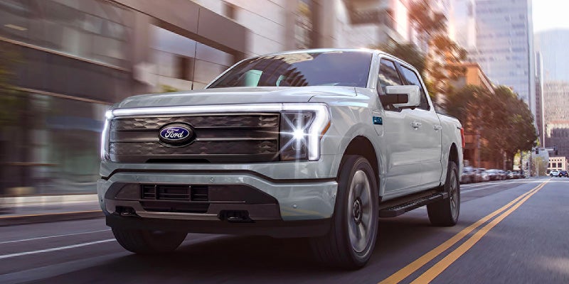 Light grey Ford F-150 Lightning electric pickup truck driving down a city street.