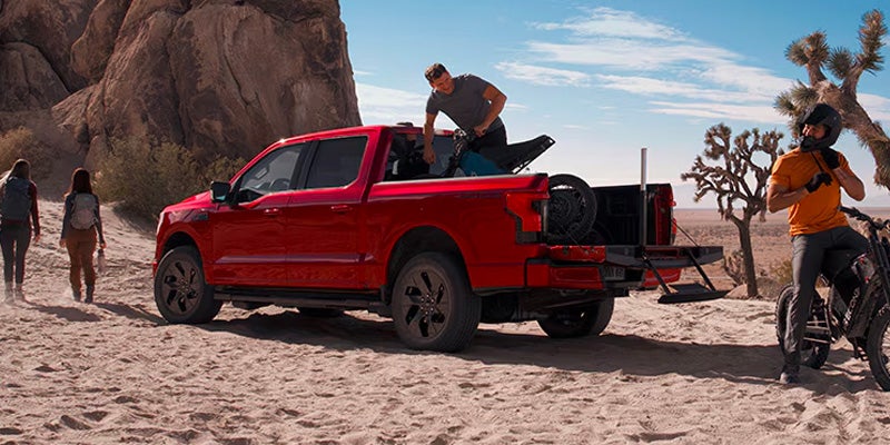 Red pickup truck in a desert with people loading gear and preparing for a ride; two hikers walk in the background.