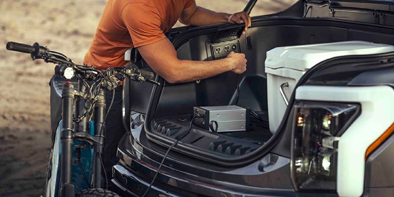 Man plugging an electric bike charger into outlets in the back of an electric truck, with a cooler beside it.