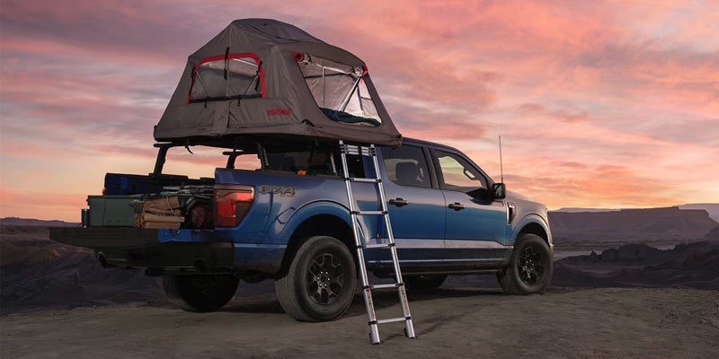 Blue pickup truck with an open rooftop tent and ladder, filled with camping gear, parked in a desert at sunset.