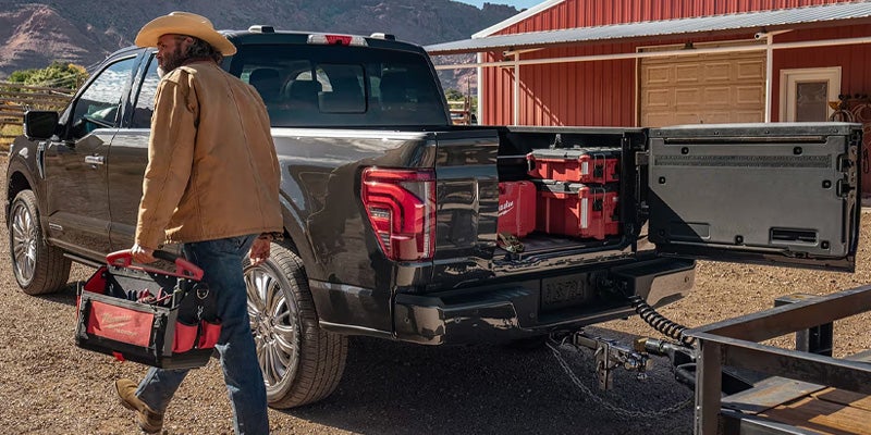 Man with cowboy hat and toolbox walks from a dark pickup truck with an open tailgate, revealing red tool cases inside.