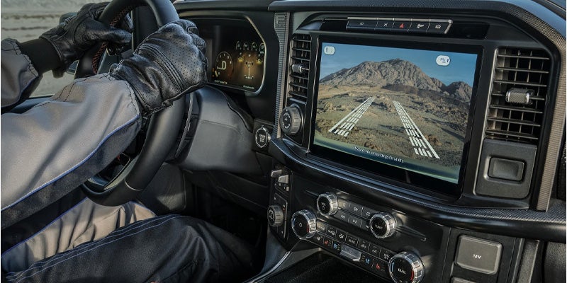 Driver in driving gloves, dashboard with a large screen displaying a backup camera view of rocky terrain with guiding lines.