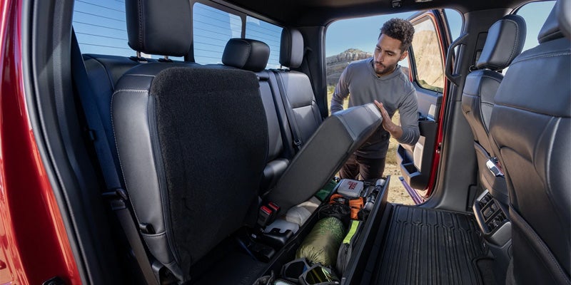Man lifts a rear truck seat revealing an under-seat storage compartment filled with outdoor gear.