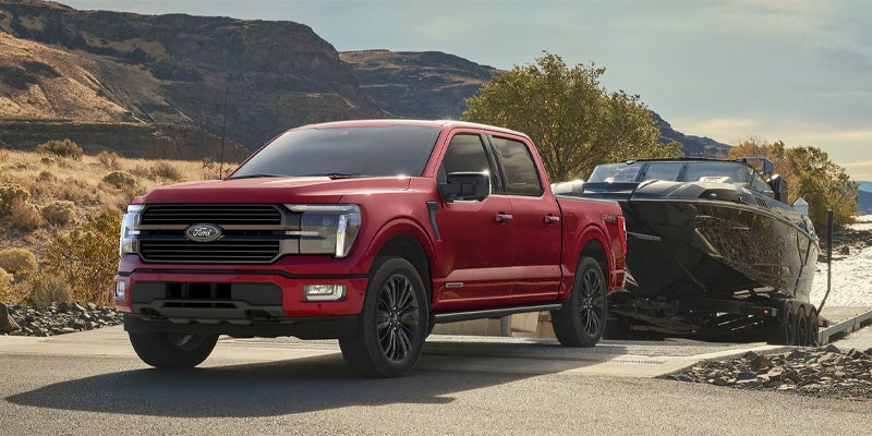 Red Ford F-150 truck towing a black boat on a trailer down a ramp, with mountains and water in the background.