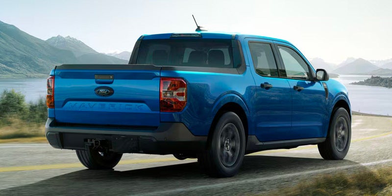 Blue Ford Maverick pickup truck driving away on a winding road with mountains and water in the background.