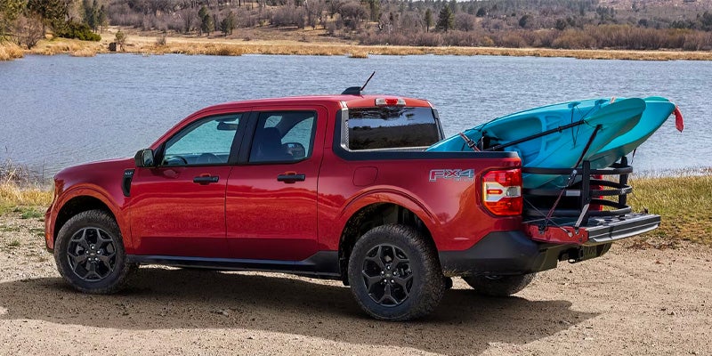 Red pickup truck with a blue kayak in its bed, tailgate down, parked by a lake on a dirt path.