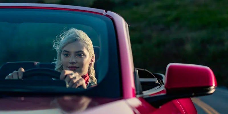 Blonde woman with windblown hair driving a red convertible car, hands on the steering wheel, looking ahead.