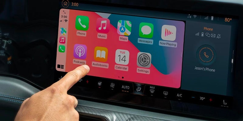 Hand touching a car's infotainment screen displaying Apple CarPlay with various app icons like Phone, Music, and Maps.