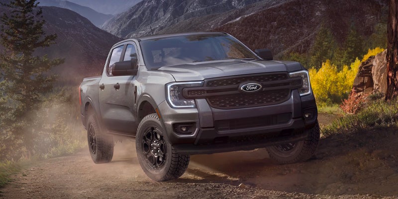 Gray Ford Ranger truck driving on a dusty mountain trail with trees and rocky hills in the background.