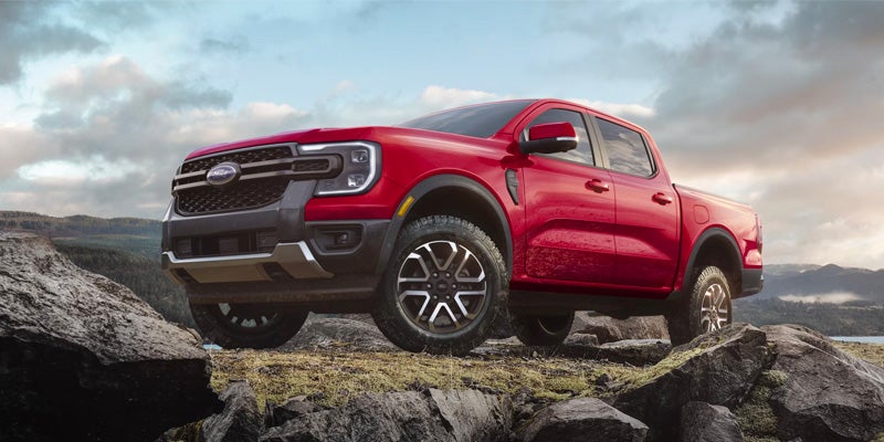 Red Ford Ranger pickup truck with a black grille and silver accents, parked on rocky terrain with mountains and a cloudy sky.