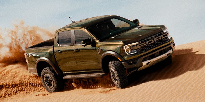 Olive green Ford Ranger Raptor pickup truck driving dynamically through desert sand dunes, kicking up a cloud of dust.