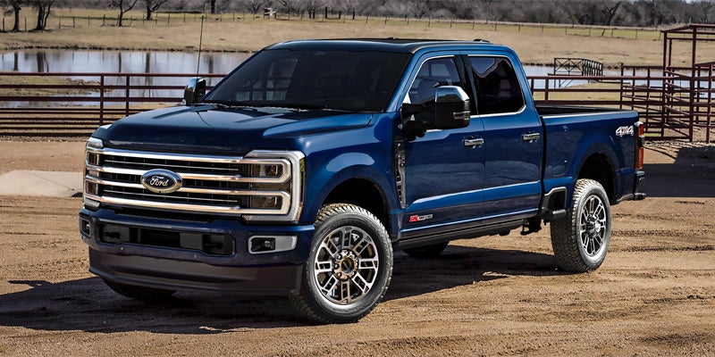 Dark blue Ford F-Series Super Duty pickup truck parked on dirt with a pond and fence in the background.