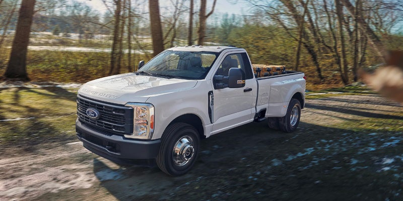 White Ford Super Duty Dually pickup truck with toolboxes in bed, driving on a dirt road through trees.