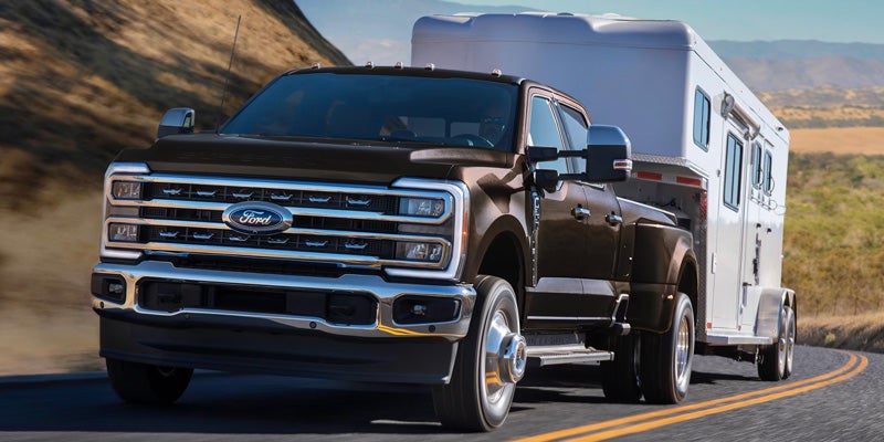 Dark brown Ford dually truck towing a white trailer on a curved road with hills in the background.