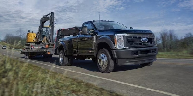 Black Ford pickup truck towing a yellow excavator on a flatbed trailer down a road.