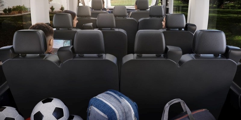 Interior view of a vehicle with multiple rows of black seats, several passengers, soccer balls, and bags.