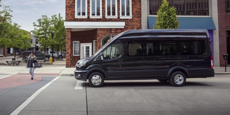 Black passenger van parked on a city street in front of brick buildings, with a person walking on the sidewalk.